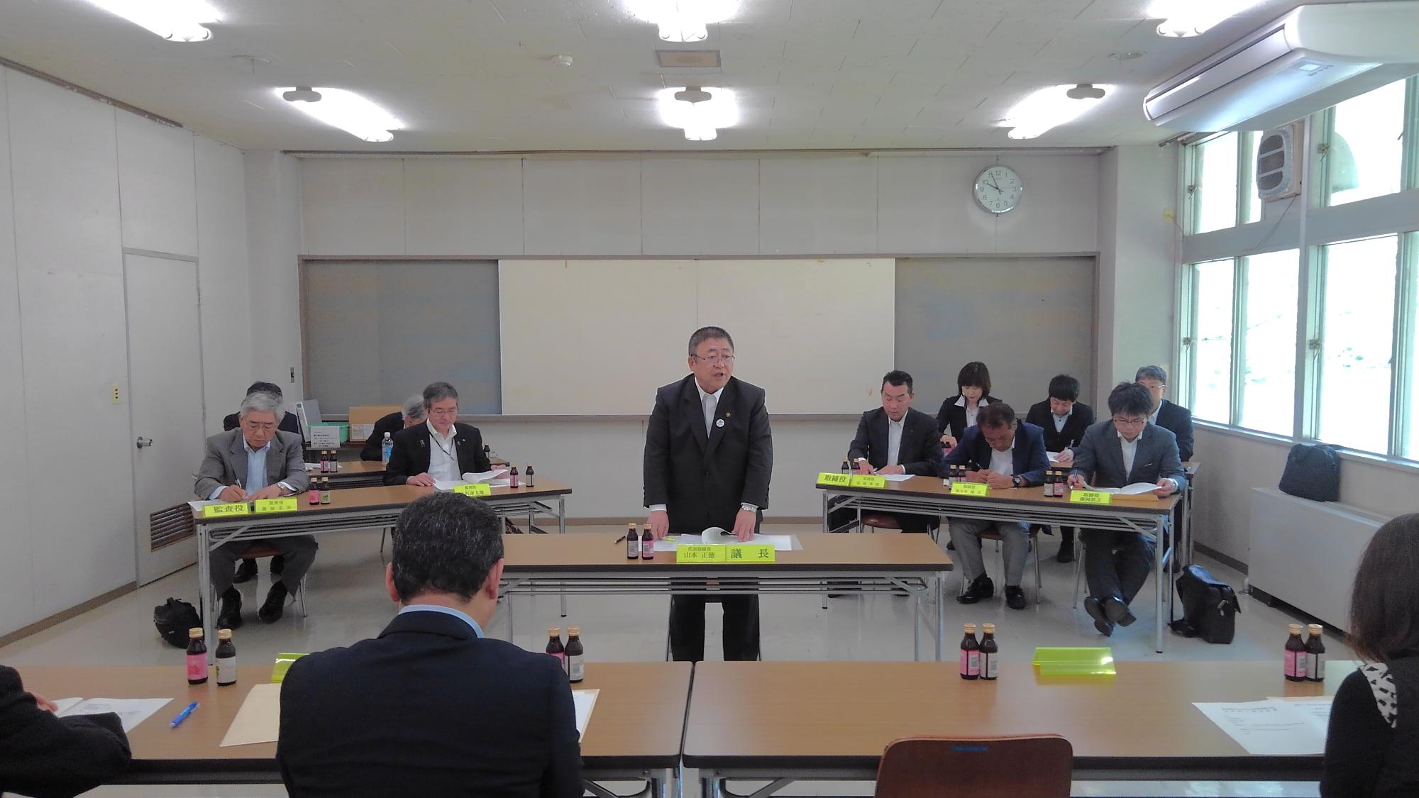 ハの字に並べられた長机の席に着く関係者と、その中央の少し前に置かれた机に立ち話をする様子の市長の写真
