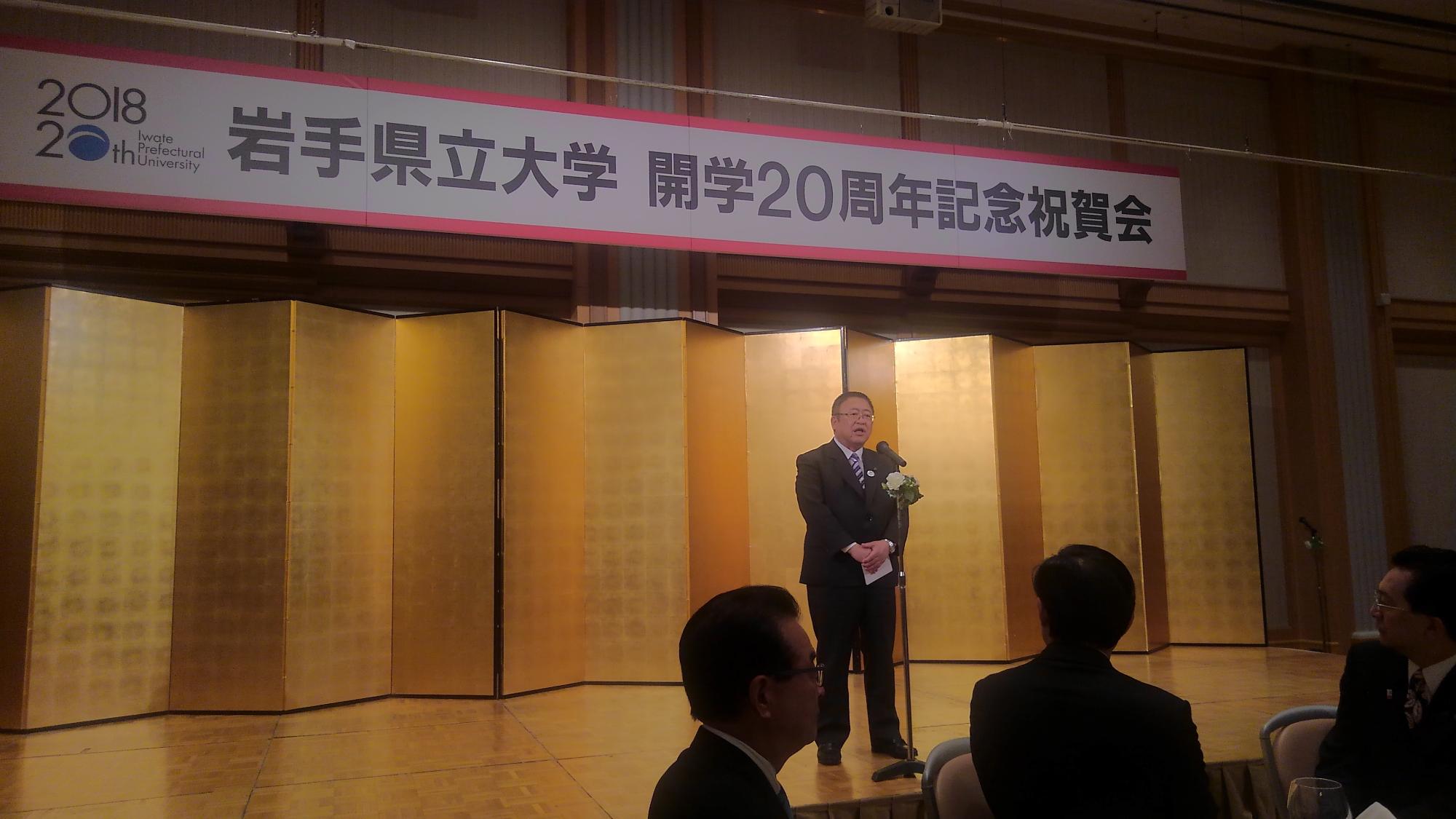 「岩手県立大学 開学20周年記念祝賀会」と書かれたパネルが吊るされ、金屏風が置かれた舞台上で、スタンドマイクに向かい話をする様子の市長の写真