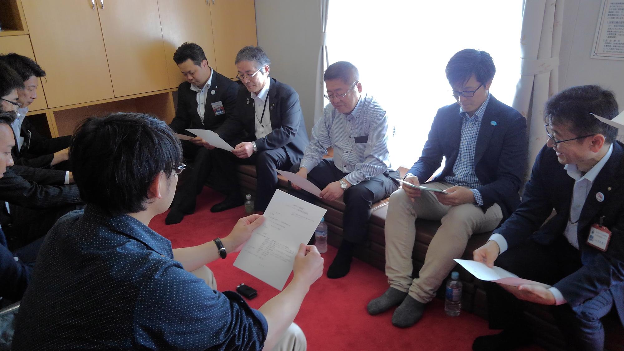 並べられた椅子に向かい合うように座り、両手で持った資料を見る市長と関係者の方々の写真