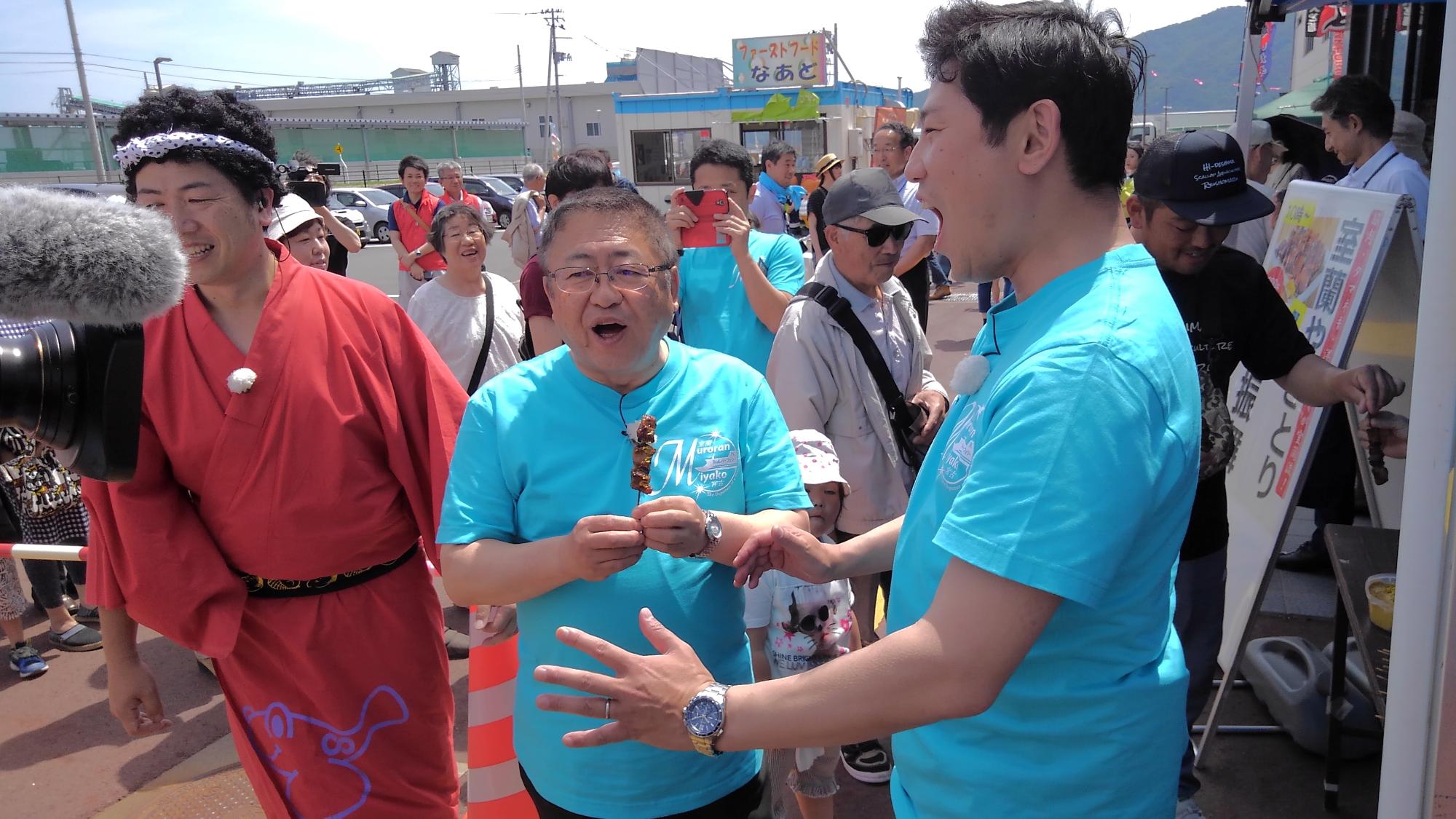青いTシャツを着た男性と、串焼きを食べている市長、赤い着物を着て頭に鉢巻を巻いた男性が写っている写真
