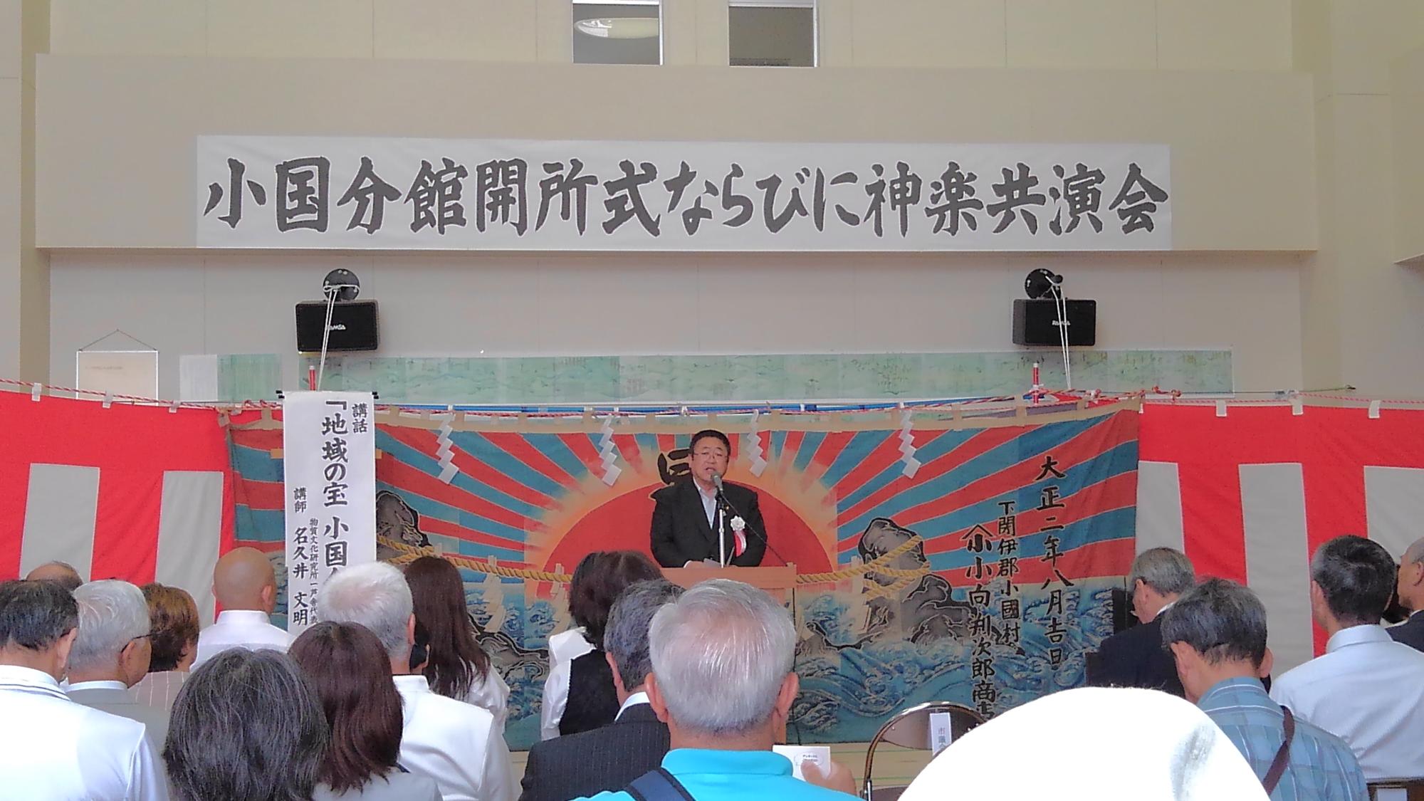 「小国分館閉所式ならびに神楽共演会」書かれた横断幕がと紅白幕や海と太陽の絵が描かれた幕が掛けられた壁の前で、マイクに向かい話をする様子の市長と椅子に座り整列して話を聞く様子の参加者の写真
