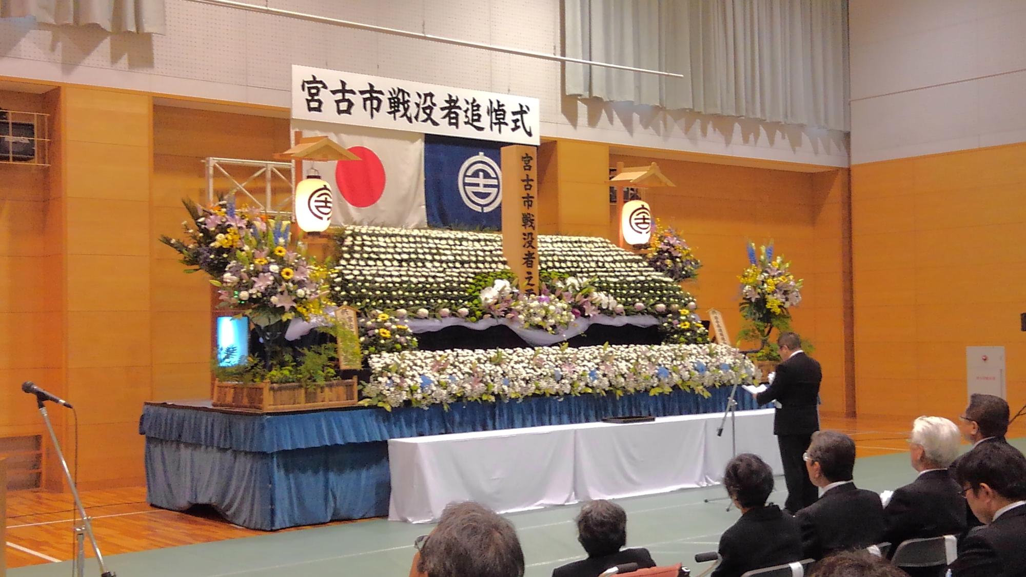 「宮古市戦没者追悼式」と書かれたボードと日本国旗、宮古市の市町村旗が吊るされ、花がたくさん並べられた献花台の前に置かれたスタンドマイクに向かい話をする様子の市長の写真