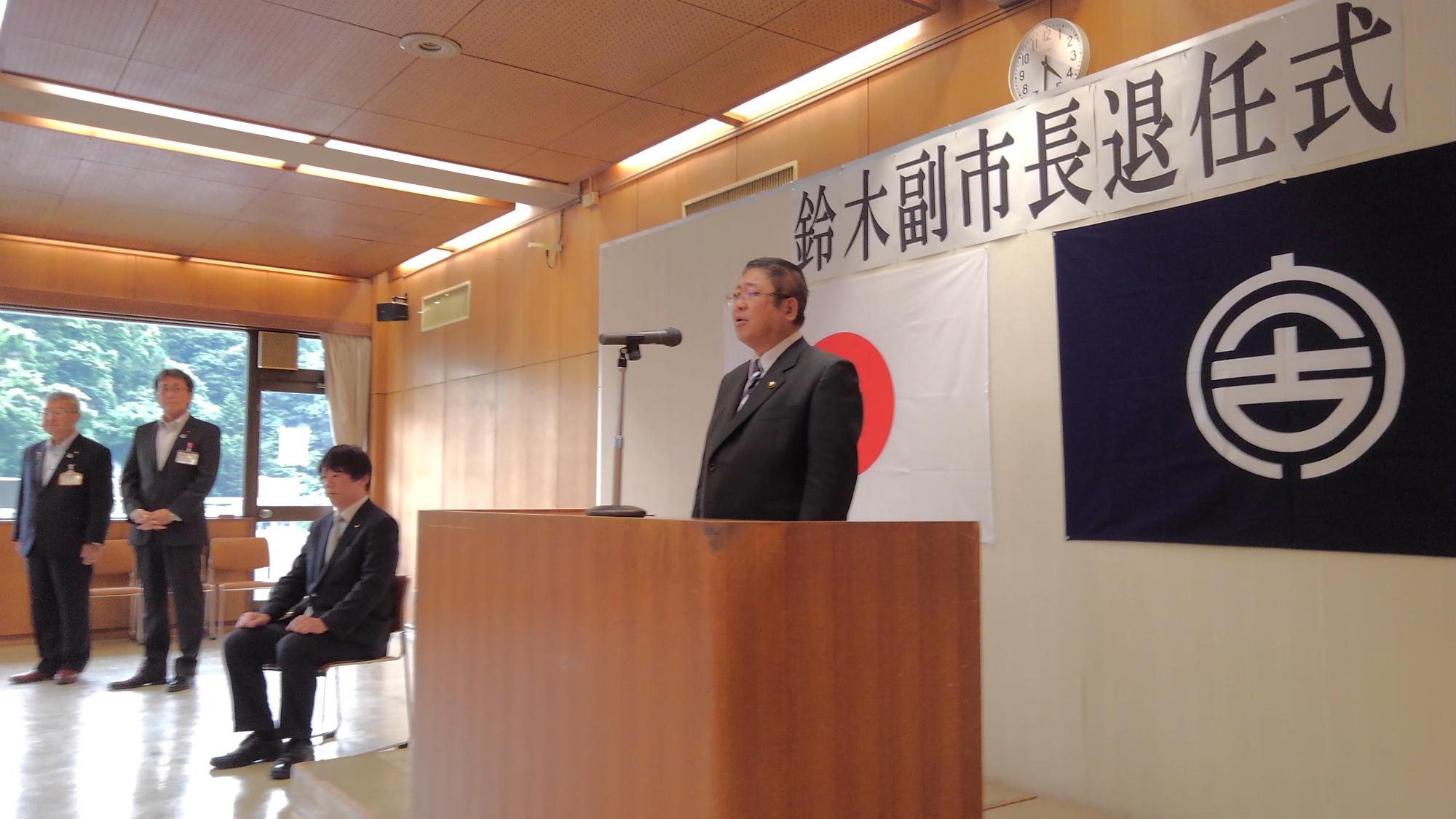 「鈴木副市長退任式」と書かれた紙と日本国旗、宮古市の市町村旗が貼られたボードの前に置かれたマイクに向かい話をする様子の市長の写真