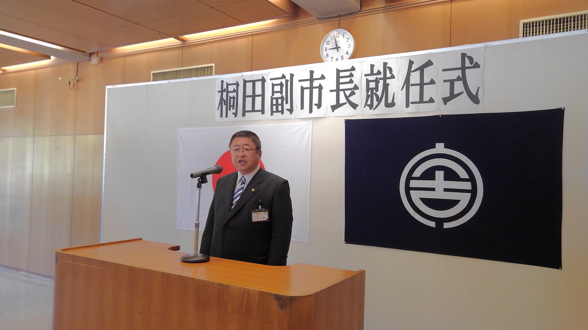 「桐田副市長就任式」と書かれた紙と日本国旗、宮古市の市町村旗が貼られたボードの前に置かれた演台で話をする様子の市長の写真