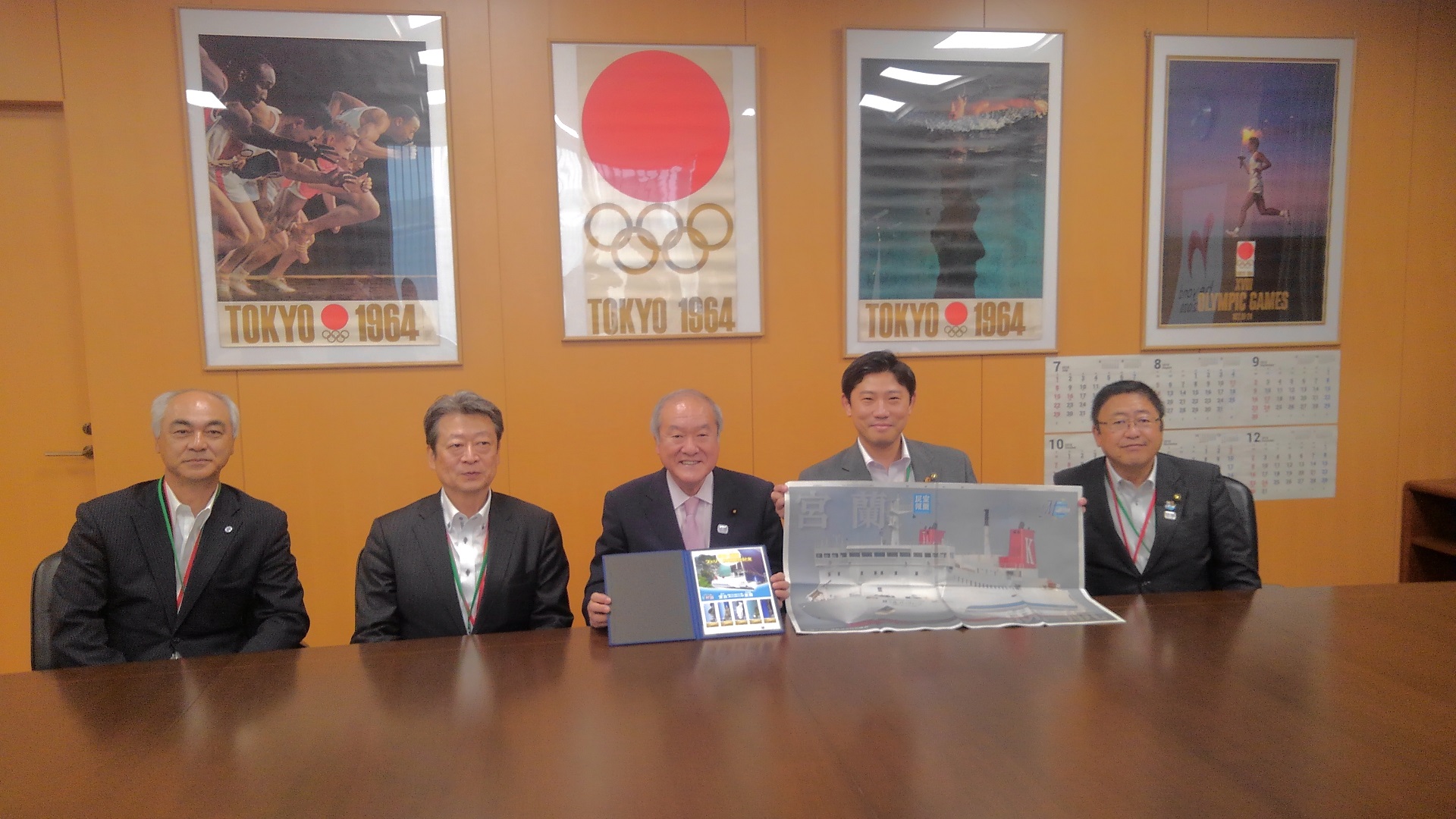 オリンピックのポスターが4枚掛けられた壁の前のテーブルの席に着き、記念撮影をする市長と4名の関係者の男性の写真