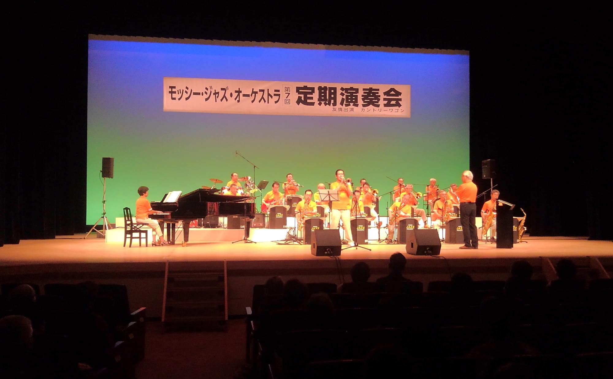 「モッシー・ジャズ・オーケストラ第7回定期演奏会」と書かれた横断幕が吊るされた舞台上でピアノや楽器を演奏する様子の方々の写真