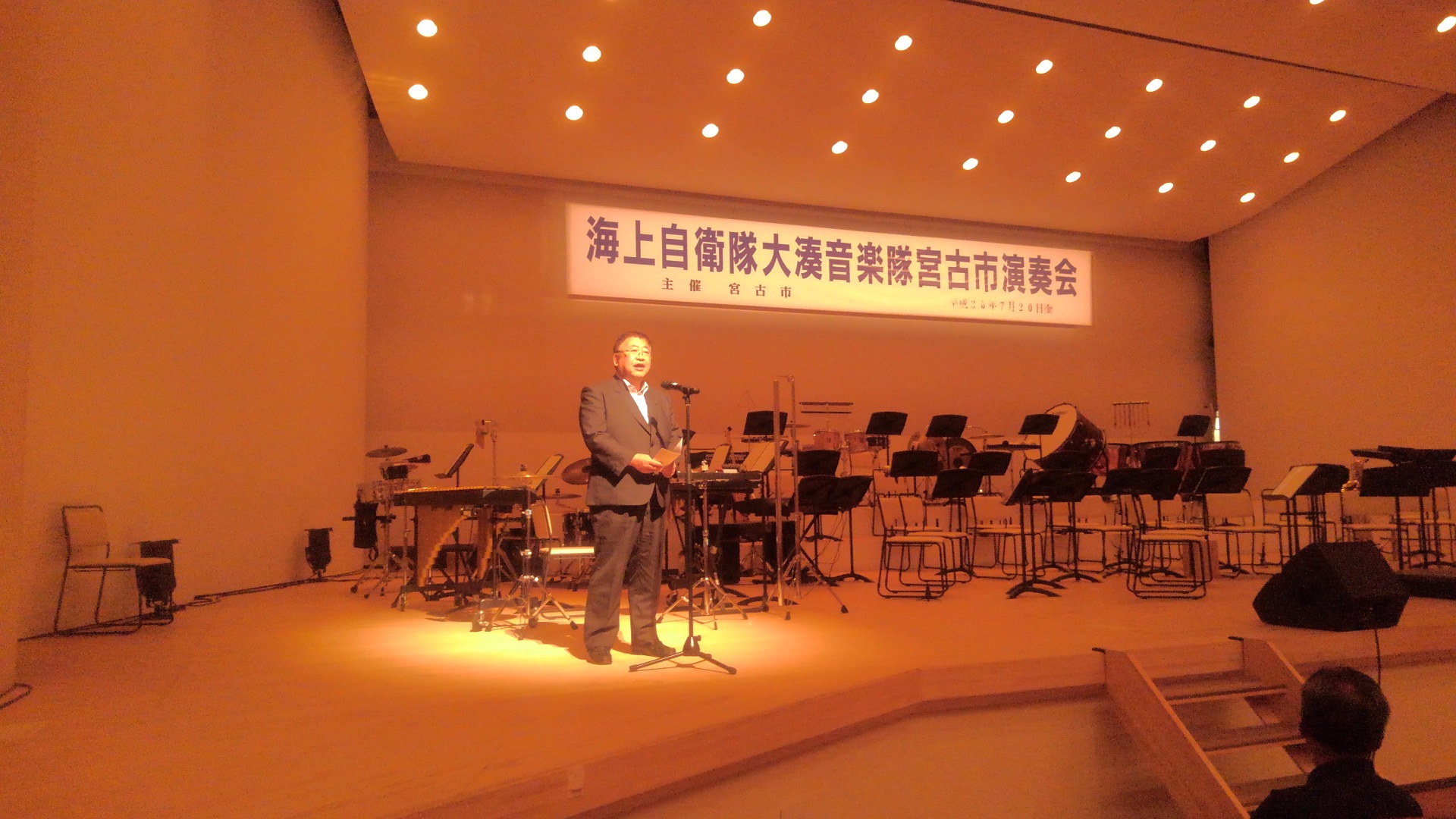 「海上自衛隊大湊音楽隊宮古市演奏会」と書かれた横断幕が設置され、楽器や譜面台が置かれた舞台上で、ライトアップされる中スタンドマイクに向かい話をする様子の市長の写真