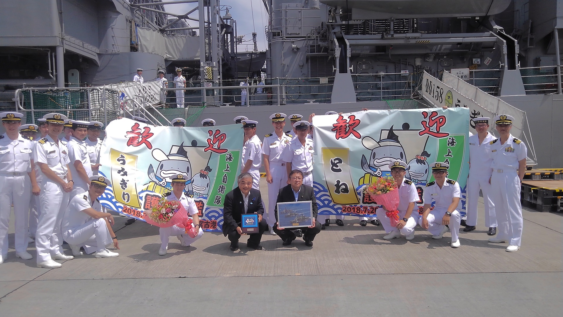 「歓迎」という文字とサーモンくんとみやこちゃんの絵や海上自衛隊護衛艦の名前である「うみぎり」「とね」と書かれた旗を持つ白い制服を着た海上自衛隊の方々の中心でしゃがんで額縁を持つ市長と男性の記念写真