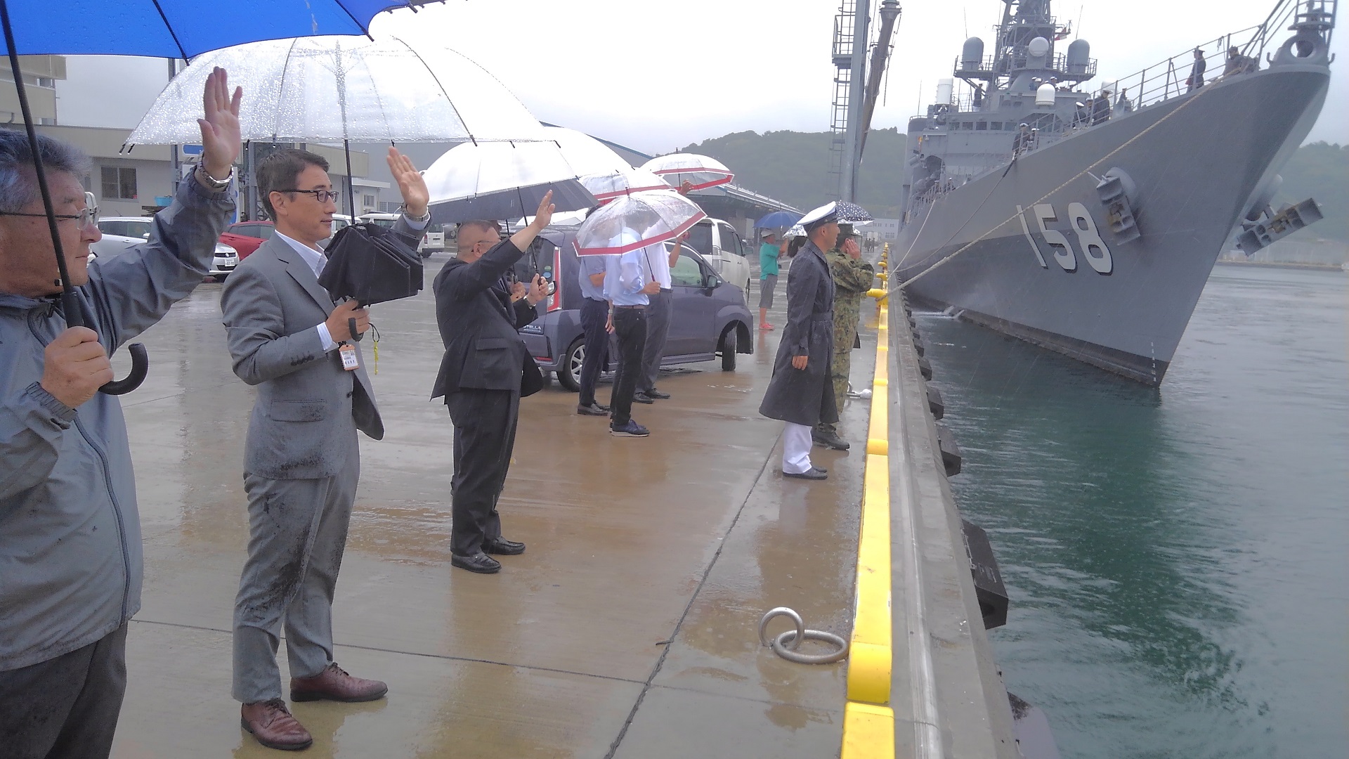 雨の降る中、傘を差し、出航する海上自衛隊護衛艦に向かって手を振る市長と関係者の方々の写真