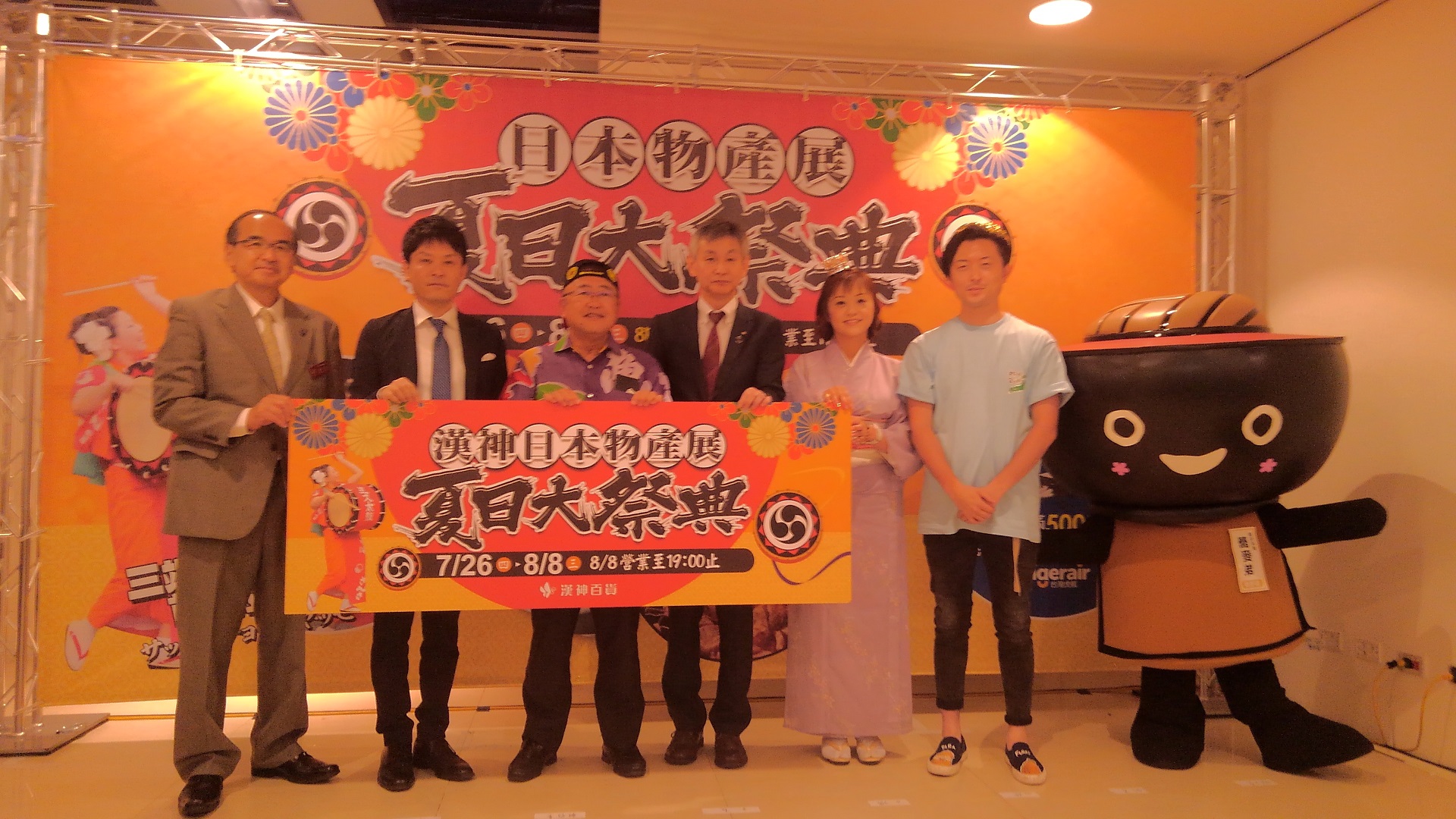 「夏日大祭典」と書かれた看板を一緒に持つ市長と4名の関係者の隣に立つ男性とわんこきょうだいの写真