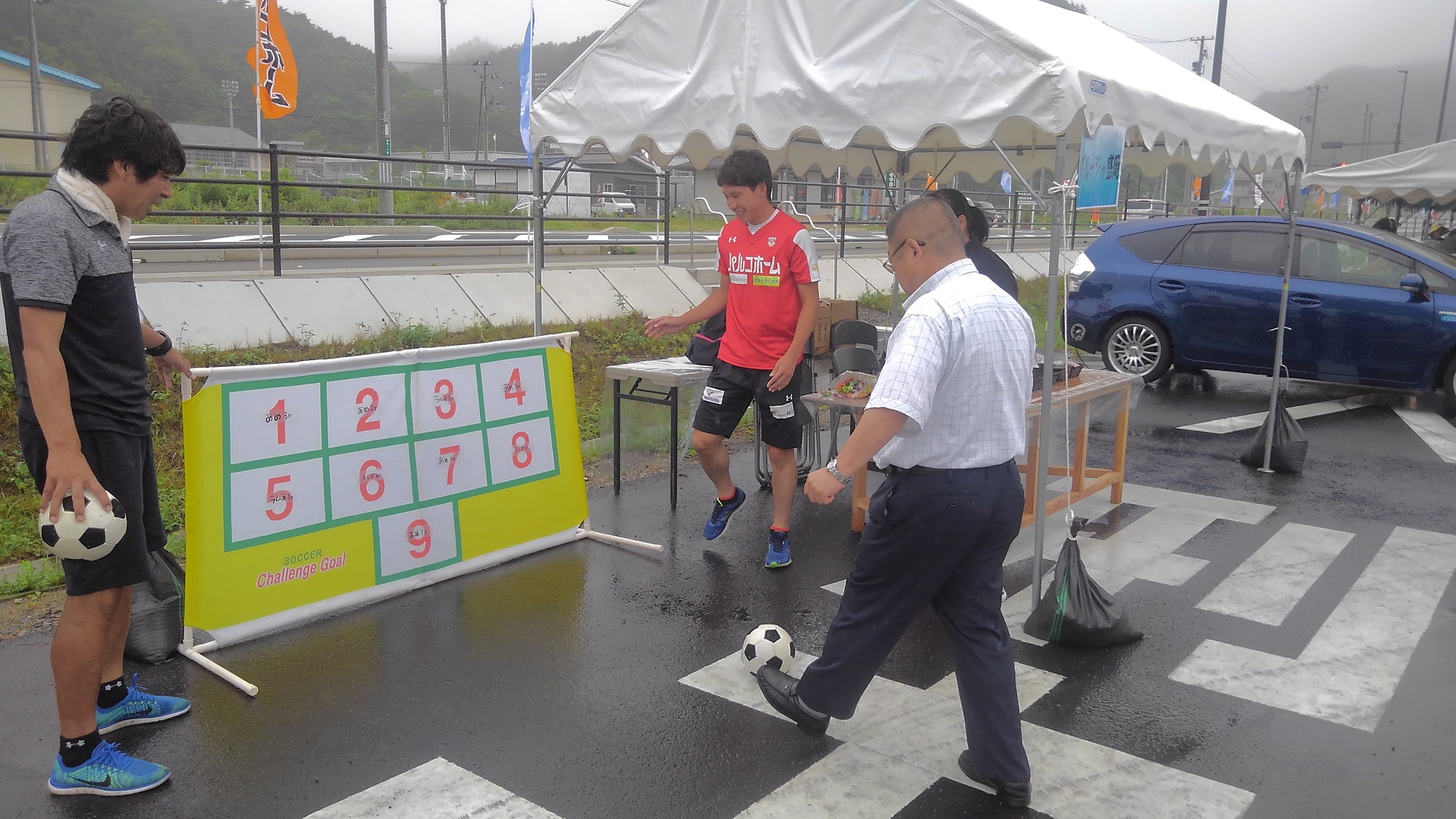 1から9までの番号が書かれたパネルに向かって、小さなサッカーボールを蹴ろうとしている様子の市長の写真