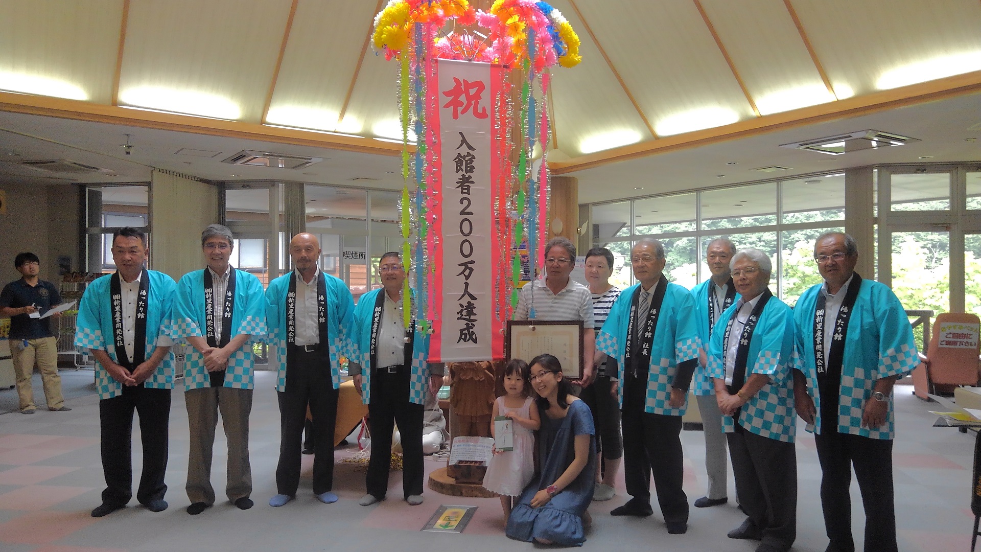 「祝 入館者200万人達成」と書かれた垂れ幕の横で、記念品や賞状を持つ家族と、家族の左右に並ぶ青い法被を着た市長と関係者7名を写した記念写真