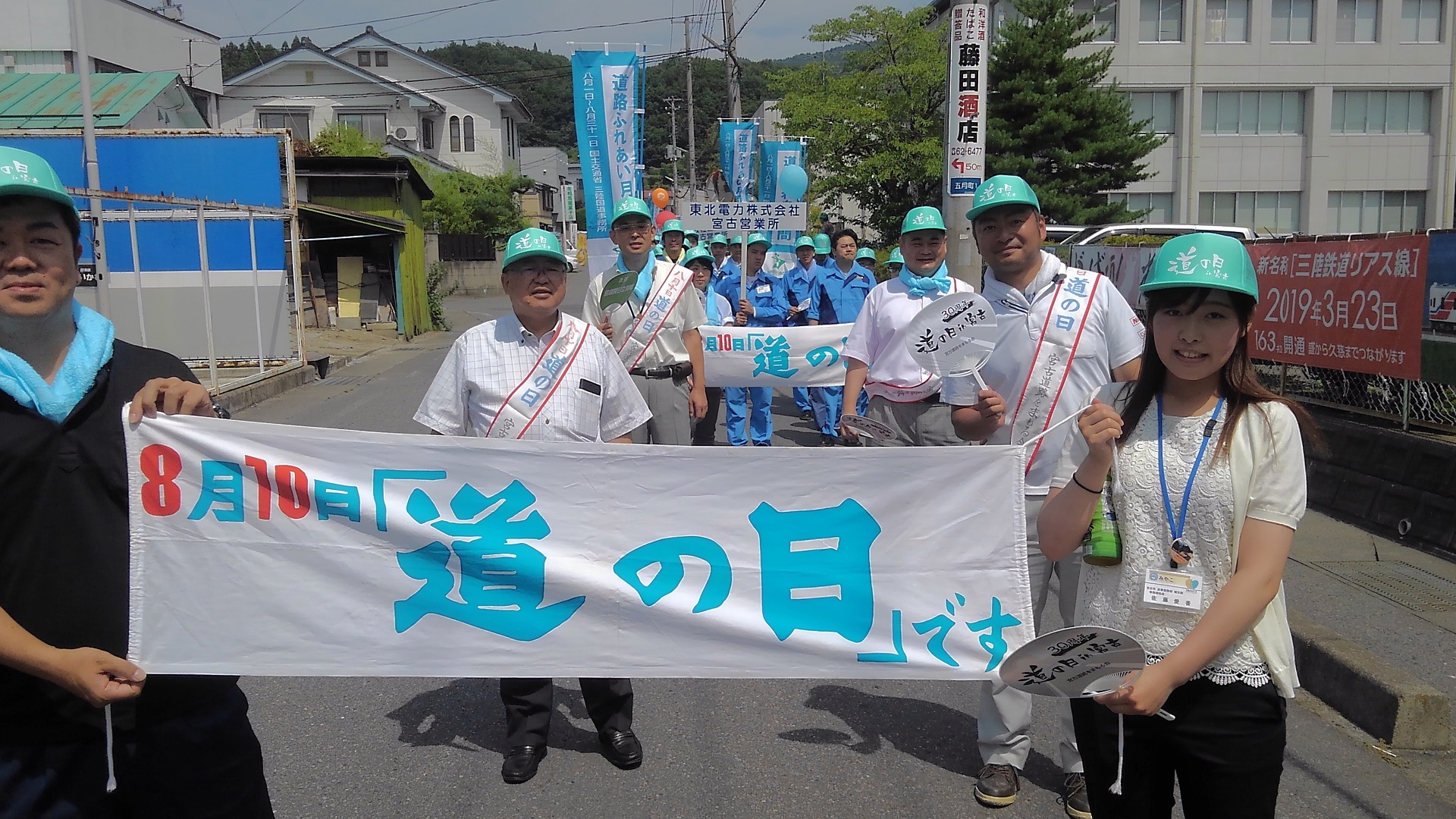 「8月10日「道の日」です」と書かれた横断幕を一緒に持つ男性と女性を先頭に、全員が青い帽子を被って並んで道路を歩く様子の写真