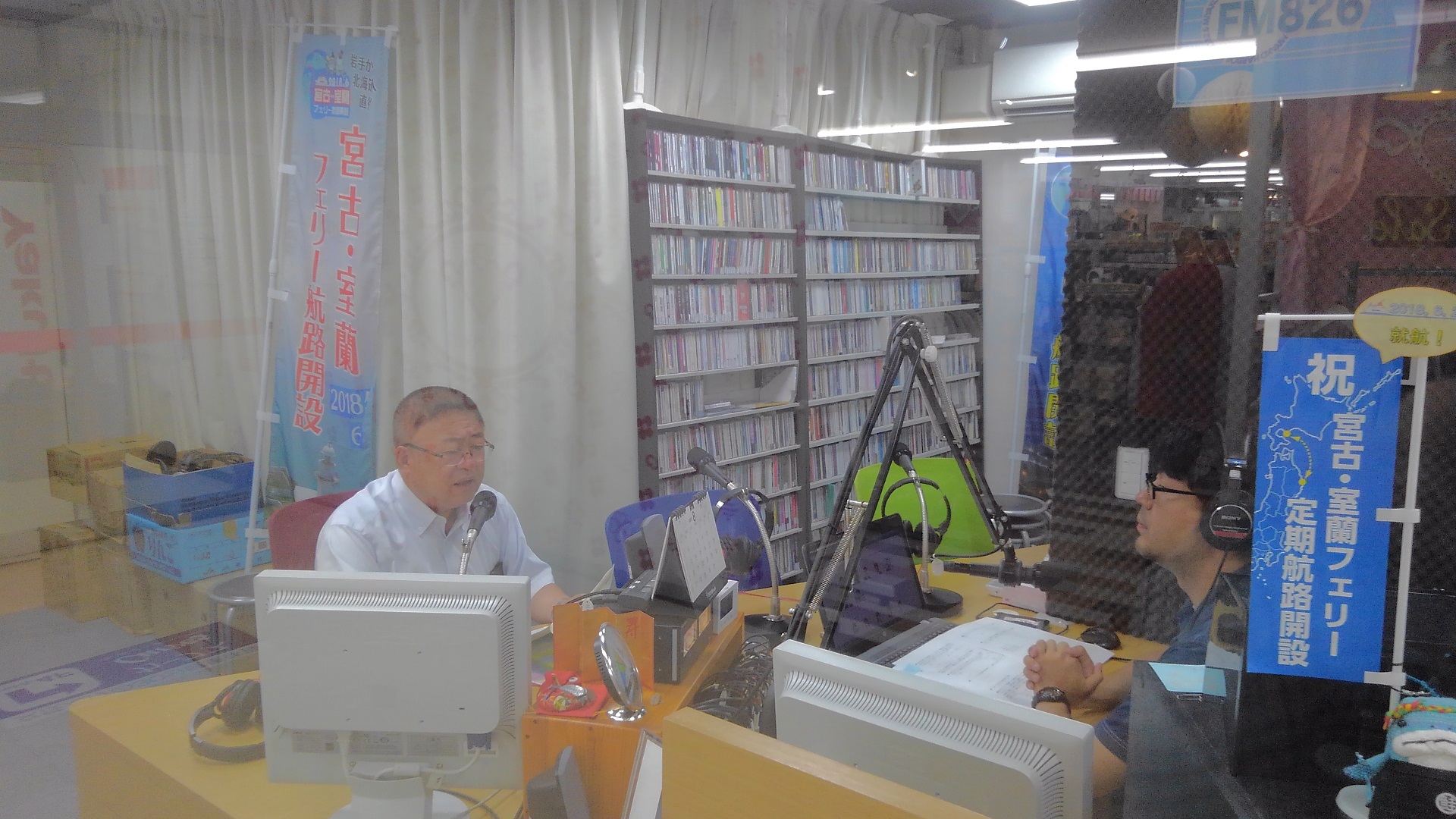 CDが並べられた棚が置かれたラジオブースで、ラジオに出演する様子の市長とパーソナリティの男性の写真