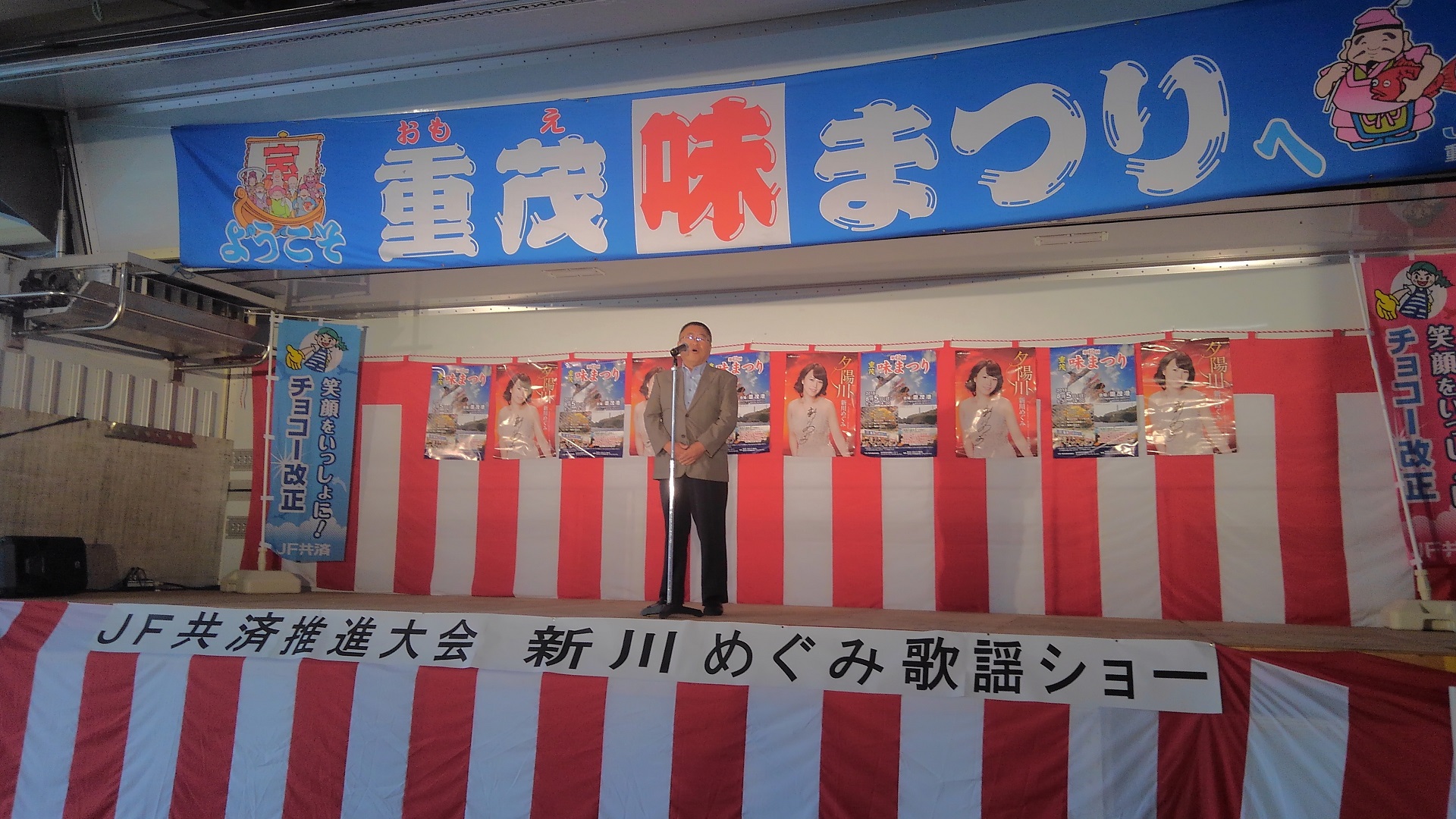 「重茂味まつり」と書かれた横断幕やポスター、紅白幕が掛けられた舞台上で、スタンドマイクに向かい話をする様子の市長の写真