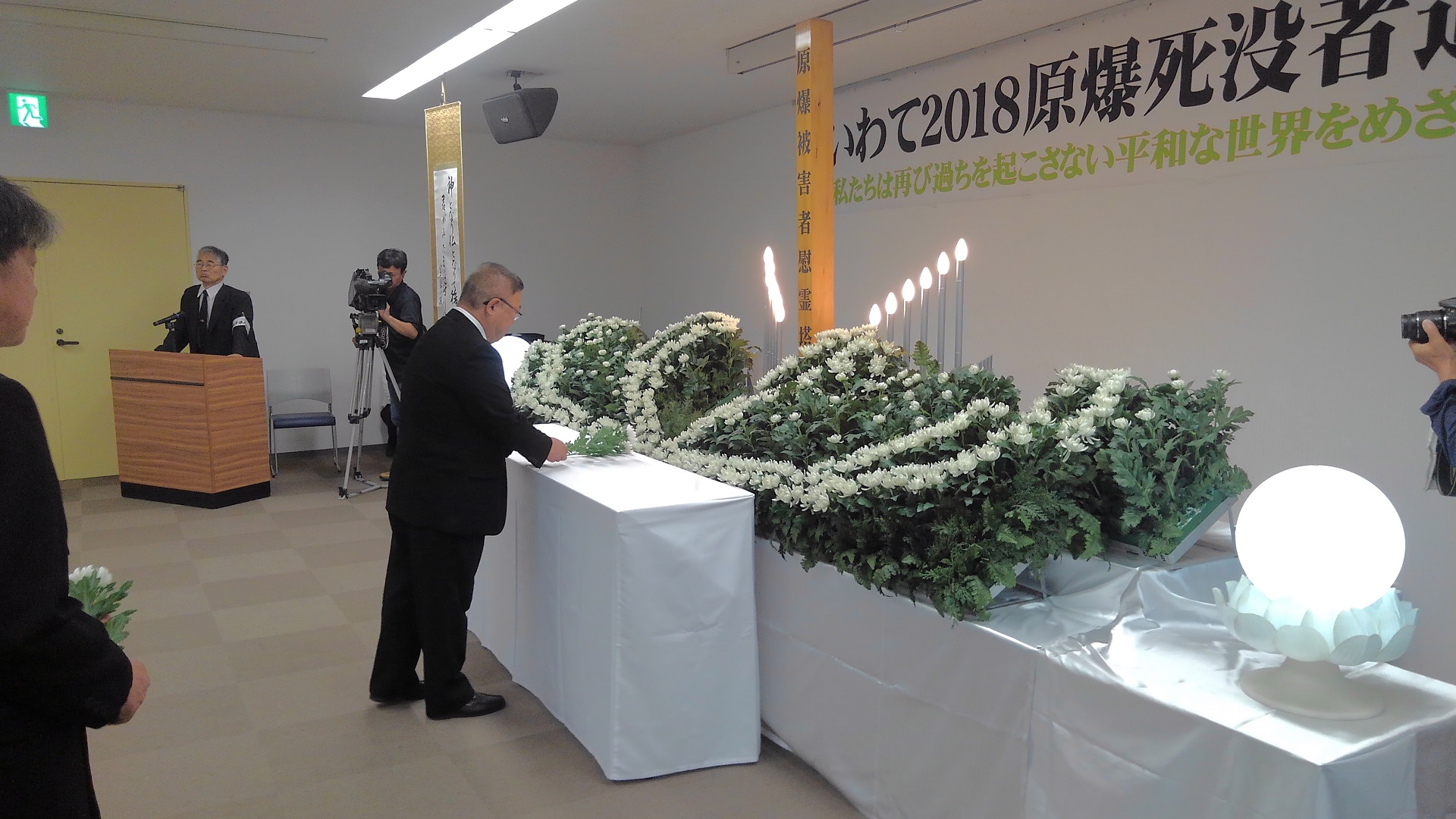 ろうそくや花が並べられた祭壇の前に置かれたテーブルに、白い花を備える様子の市長の写真