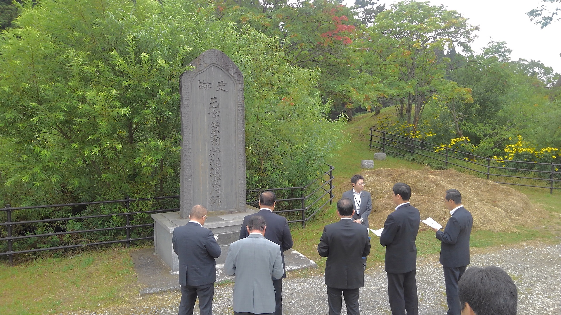 大きな縦長の楕円形の石碑の前で、資料を見ながら話を聞く様子の市長と関係者の男性5名のを後方から写した写真