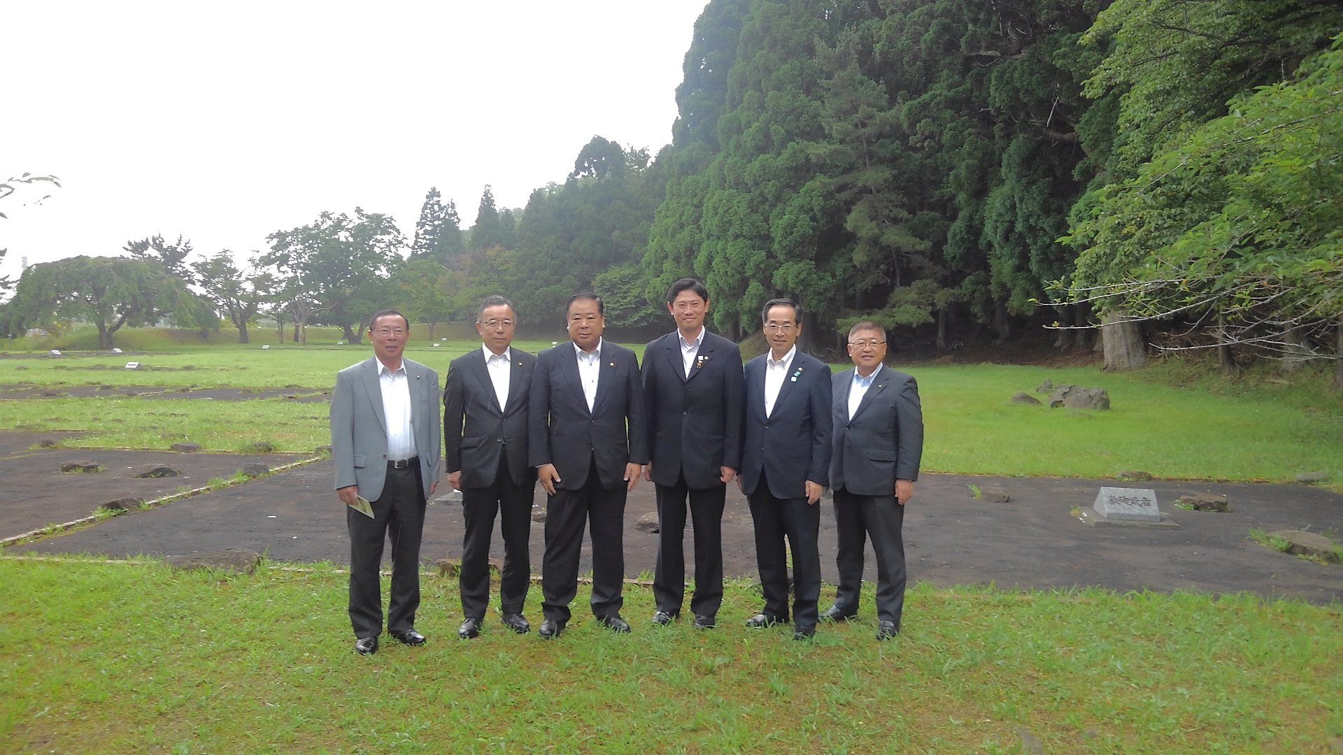 後方に大きな木がそびえたつ芝生の広場で、市長と関係者の男性5名が横一列に並んでいる記念写真