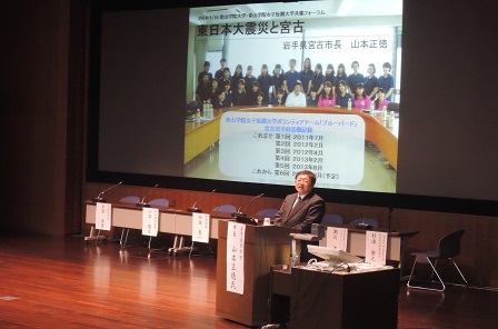 画面が映されたスクリーンの斜め前に置かれた演台に向かい、講演する様子の市長の写真
