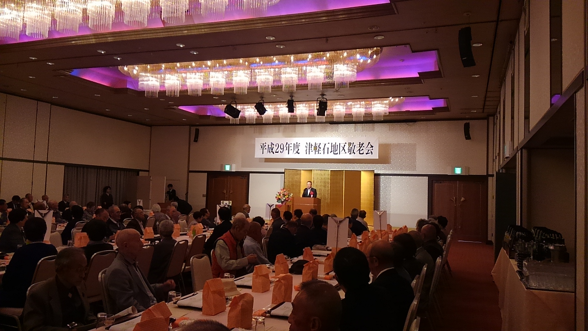 「平成29年度 津軽石地区敬老会」と書かれた横断幕が掲げられ金屛風が設置された檀上に胸章リボンを付けた市長が立ち、自席に座った参加者が話を聞いている敬老会の様子の写真